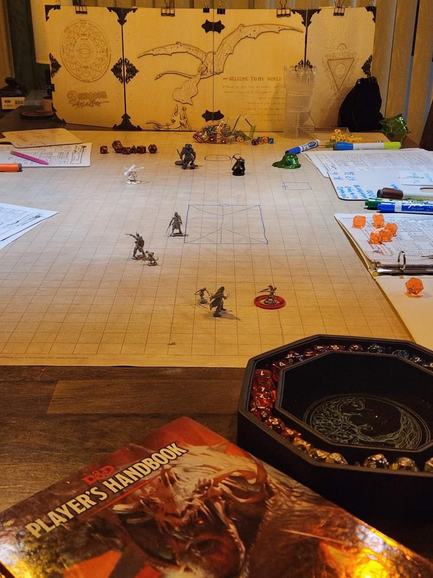 a table arranged to play dungeons and dragons with a map, dice, and game figures sitting on it. a wooden game screen sits at the end of the table with a dragon emblem