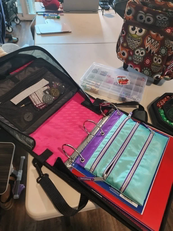 an open 3 ring binder on a table. inside is a pouch, notebook, and folders. Also on the table are a storage container and backpack.