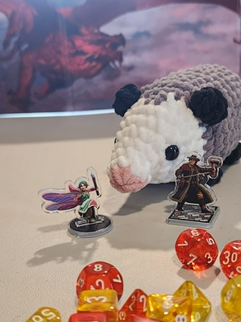 Miniature roleplayer stand in figures next to a crocheted possum with orange and yellow dice